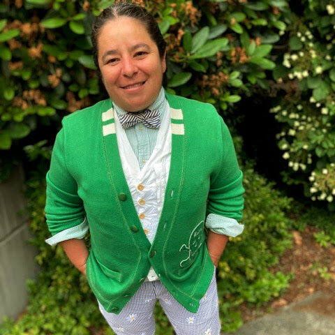 Nadine Maestas standing in front of lush greenery with a matching green sweater, floral pants, and black and white striped bowtie.