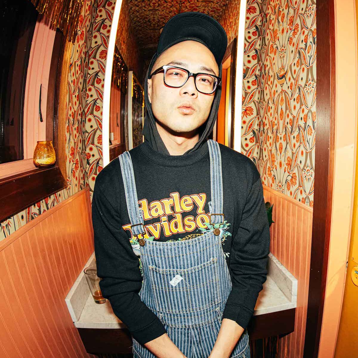 Richard Chiem wearing glasses, a baseball cap, overalls, and a Harley Davidson hoodie. He is posed in front of a hallway with patterned wallpaper.